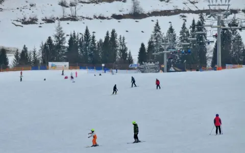 Narciarze i snowboardziści szusują po ośnieżonym stoku w Bukowinie Tatrzańskiej. W tle widać wyciąg krzesełkowy i las.