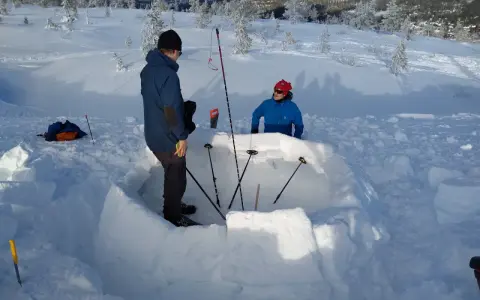 Två personer bygger en bivack i snön. De använder snöblock för att bygga väggarna och stavar för att markera utrymmet.