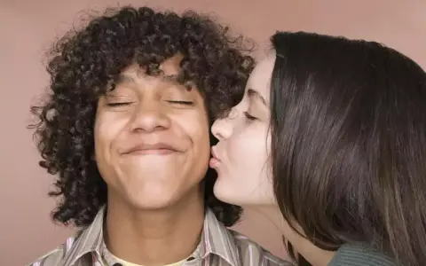 Un jeune homme aux cheveux bouclés reçoit un baiser sur la joue d'une jeune femme. Il semble ravi, les yeux fermés. Un moment de tendresse, un vrai pécho def.