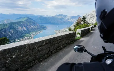 Widok z motocykla na Zatokę Kotorską w Czarnogórze. Kręta droga z kamiennym murem i zapierająca dech w piersiach panorama gór i morza.