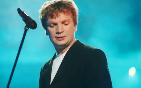 Grzegorz Ciechowski, w czarnym garniturze, pochyla się nad mikrofonem. Jego niezapomniane utwory wciąż brzmią w sercach fanów.