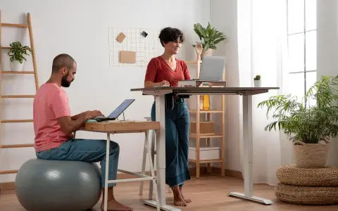 Un homme travaille sur son ordinateur, assis sur un ballon de gym, tandis qu'une femme travaille debout à un bureau réglable. Un espace de travail moderne et dynamique.