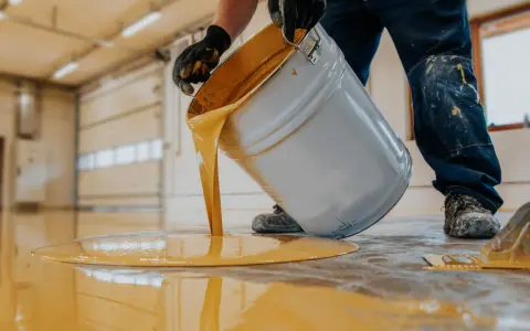Persona con guantes vierte pintura amarilla de un cubo para pintar suelo cemento sin pulir.