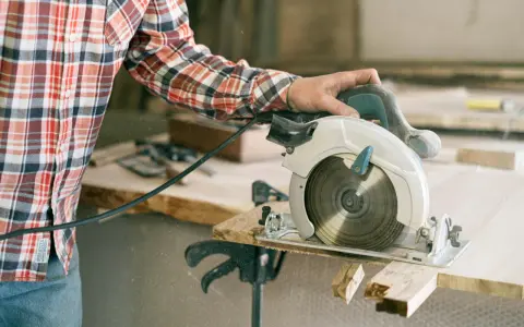 A person in a plaid shirt demonstrates how to use a circular saw, cutting wood with sawdust flying.