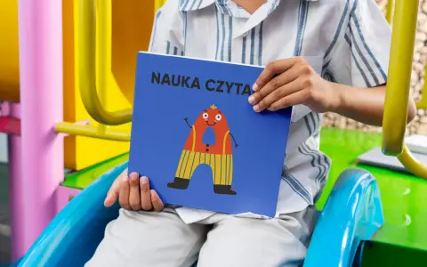 Chłopiec trzyma książkę "Nauka czytania dla 6 latka" z zabawną literką "A" na okładce.
