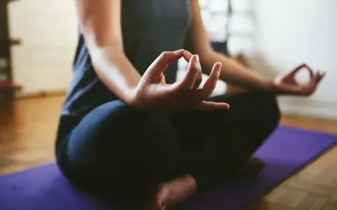 En person sitter i meditationsställning på en lila matta. Händerna vilar i knät med tumme och pekfinger sammanfogade, en vanlig gest för att visa hur man mediterar.