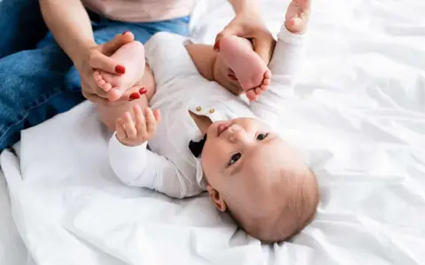 Mama delikatnie ćwiczy nóżki niemowlaka, pomagając mu przy wzmożonym napięciu mięśniowym. Dziecko leży na białym tle.