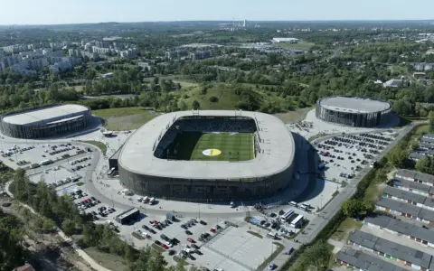 Widok z lotu ptaka na nowoczesny kompleks sportowy, w tym stadion piłkarski i dwie hale widowiskowe. To arena Sosnowiec, otoczona zielenią i parkingami.