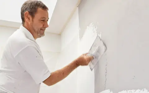 Mann verputzt eine Wand mit einer Kelle. Die Arbeit im Bad ist fast fertig.