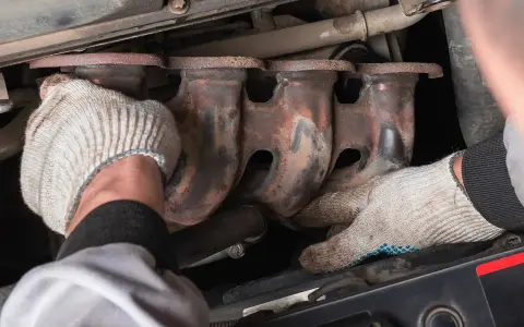 Mechanic's gloved hands work on a rusty exhaust manifold, possibly to fix an engine ticking noise.