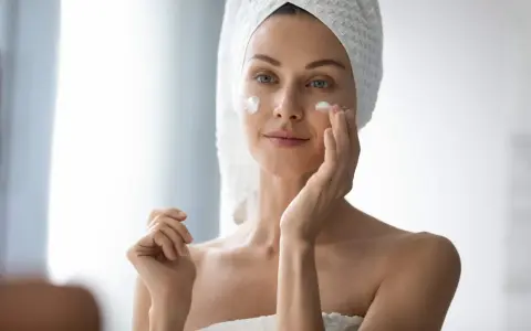 Femme appliquant de la crème sur son visage pour lutter contre la xerose cutanée. Elle a une serviette sur la tête.