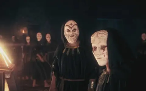 Miembros del culto de Kosmos, con túnicas oscuras y máscaras blancas, se reúnen ante un objeto piramidal brillante.