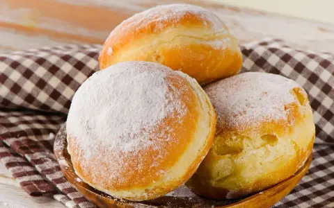 Trzy puszyste pączki, dwa posypane cukrem pudrem, leżą na drewnianej misce. Idealne na tłusty czwartek, te fit pączki kuszą apetycznym wyglądem.