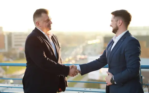 Dwóch mężczyzn w garniturach ściska sobie dłonie na tle miasta.