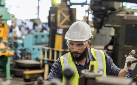 Obrero con casco y gafas de seguridad trabajando en una máquina en una fábrica, parte esencial de la manufactura moderna.