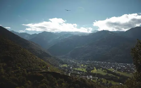 Vue aérienne d'une vallée verdoyante avec un village niché, entouré de montagnes majestueuses. Un planeur plane dans le ciel bleu, rappelant les paysages de Toulouse et ses environs.