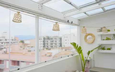 Un cerramiento ático con vistas urbanas, decorado con lámparas de mimbre y plantas.
