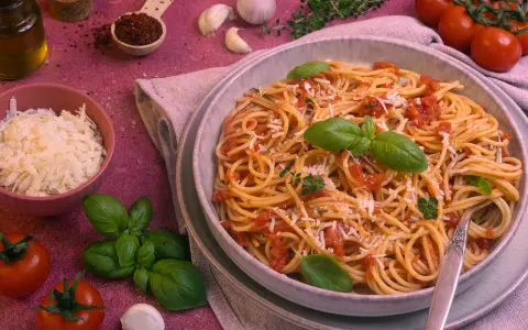 Ein Teller Spaghetti Napoli mit Tomatensauce, Basilikum und Parmesan. Daneben Zutaten wie Tomaten, Knoblauch und Olivenöl.