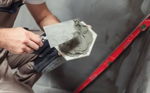 Majster nakłada klej na płytkę, przygotowując ją do ułożenia. Obok leży poziomica, a w tle widać materiał, który może być matą pod płytki na OSB.