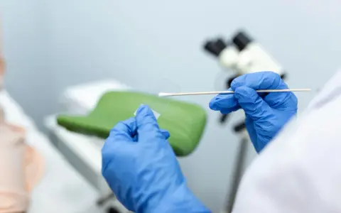 Ręce w niebieskich rękawiczkach przygotowują próbkę do badań cytologia wyniki. W tle mikroskop i fotel ginekologiczny.