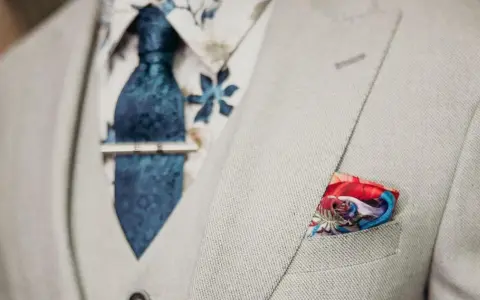 Elegancki mężczyzna w jasnej marynarce, z niebieskim krawatem i spinką do krawata jak nosić. W kieszeni kolorowa poszetka.