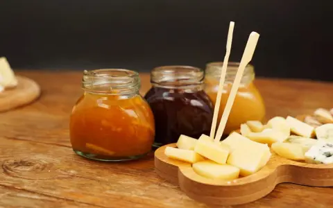 Tre vasetti di marmellata, uno arancione, uno scuro e uno giallo, accompagnano un tagliere di formaggio a cubetti, pronto per essere gustato.