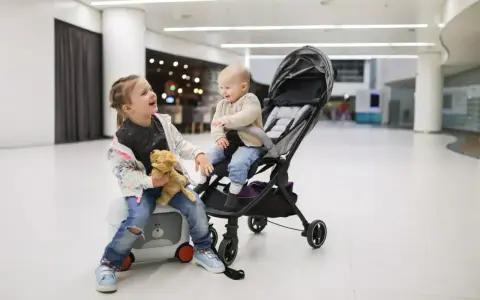 Dwie dziewczynki, jedna siedzi na walizce z misiem, druga w wózku, śmieją się w jasnym wnętrzu.