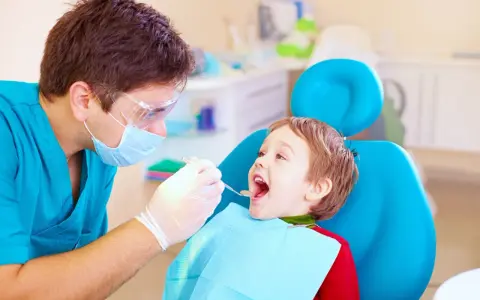 Dentysta bada uśmiechniętego chłopca, sprawdzając, kiedy najpóźniej pierwszy ząbek pojawi się u malucha.