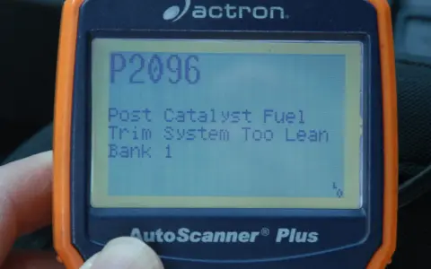 An orange and blue Actron AutoScanner Plus displays error code P2096: Post Catalyst Fuel Trim System Too Lean Bank 1.