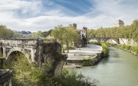 Un ponte rotto, con archi antichi e vegetazione rigogliosa, domina un fiume serpeggiante a Roma.