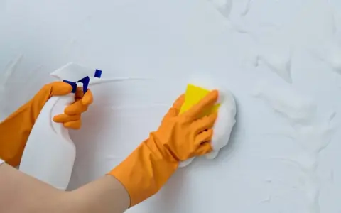 Manos con guantes naranjas limpian paredes blancas con spray y esponja. Guía práctica: como limpiar paredes blancas.