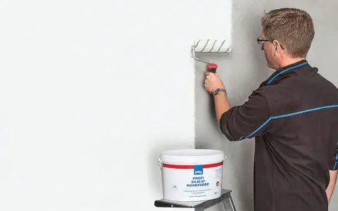 Mann streicht mit einer Farbrolle eine Wand weiß. Der Eimer mit der "Profi Silikat Wandfarbe" steht daneben. Perfekt für das Streichen bei Auszug.