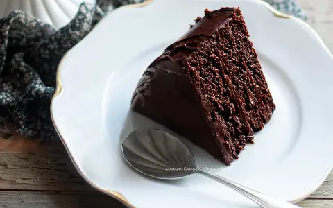 Una fetta di torta al cioccolato morbida e umida, ricoperta di glassa lucida, su un piatto bianco con un cucchiaino.