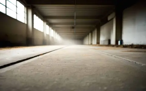 Pusta hala z betonową posadzką i rzędem okien po lewej. Długi korytarz, kurz w powietrzu. Dlaczego beton nie ma pracy?