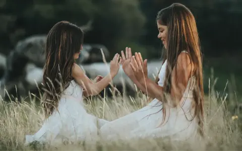 Dwie dziewczynki w białych sukienkach siedzą na łące i bawią się w "pajacyka". To idealna wyliczanka dla dzieci, która rozwija koordynację ruchową.