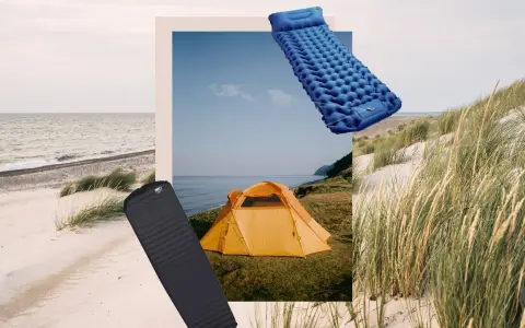 Zwei Isomatten, eine blaue aufblasbare und eine schwarze Schaumstoffmatte, liegen neben einem orangefarbenen Zelt am Strand.