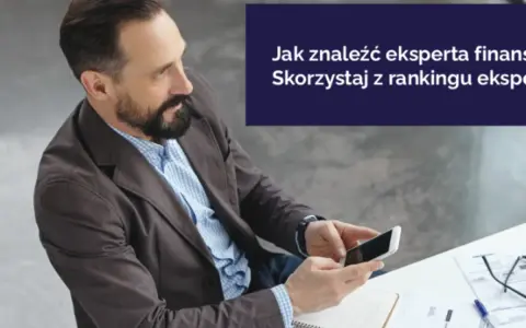 Mężczyzna z brodą trzyma smartfon, siedząc przy biurku z dokumentami.