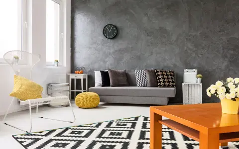 Nowoczesny salon z szarą ścianą, na której widać ciekawe efekty. Kanapa, biały fotel i geometryczny dywan tworzą przytulną przestrzeń.