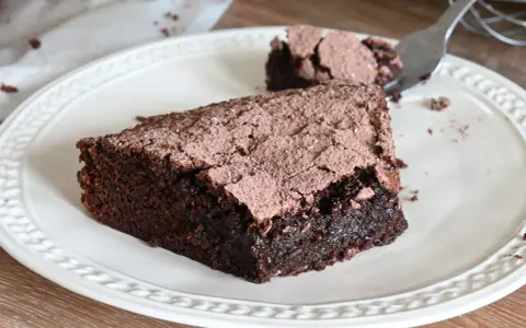 Una fetta di torta al cioccolato fondente, cosparsa di cacao, su un piatto bianco. Un pezzetto è stato staccato con una forchetta.