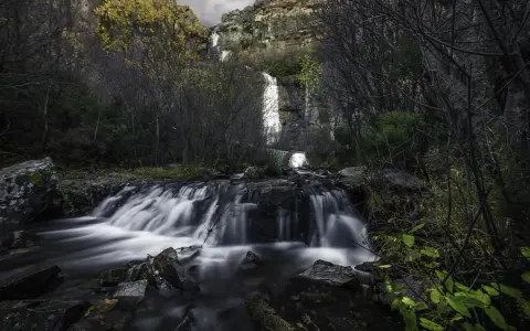 Cascada en el **río Bornova**, con aguas sedosas cayendo entre rocas y vegetación otoñal, bajo un cielo nublado.