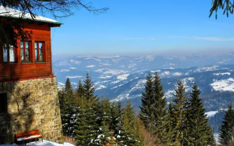 Drewniany dom na zboczu góry, z którego roztacza się widok na pokryte śniegiem pasmo górskie. W oddali widać wielki stożek górski.