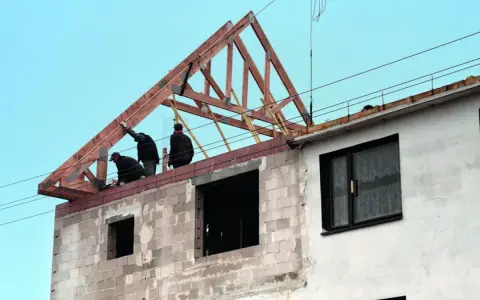 Trzech budowlańców pracuje przy podnoszeniu drewnianej konstrukcji dachu. Widać postęp prac budowlanych.