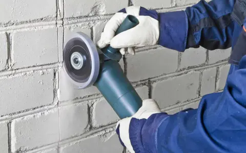 Hands in gloves use an angle grinder to cut a line into a white brick wall, demonstrating how to cut brick precisely.