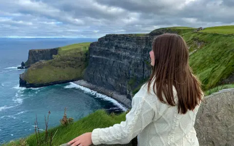 A woman in a cream cable-knit sweater gazes at the dramatic Cliffs of Moher, a breathtaking highlight of many tours of Ireland.