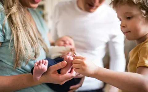 Ein älteres Kind berührt neugierig die Füßchen des Babys. Ein Hauch von eifersucht unter geschwistern liegt in der Luft, während die Eltern das neue Familienmitglied halten.