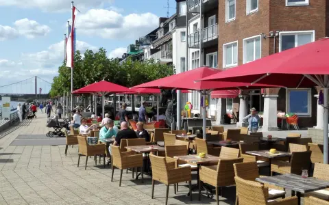 Gemütliche Straßencafés mit roten Sonnenschirmen laden zum Verweilen ein. Menschen spazieren am Rhein entlang, im Hintergrund die Brücke von Emmerich.