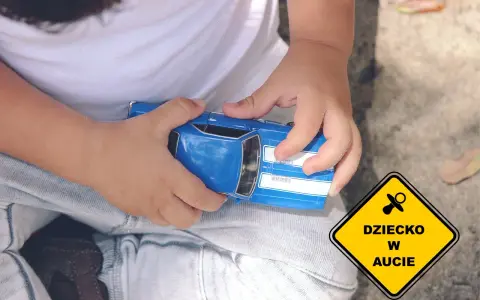 Dziecko bawi się niebieskim autkiem, a obok widnieje żółty znak "Dziecko w aucie", symbolizujący troskę o najbezpieczniejsze foteliki samochodowe.