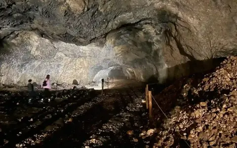 Wnętrze jaskini ciemnej, z kamienistym dnem i skalnymi ścianami. W oddali widać dwie osoby zwiedzające.