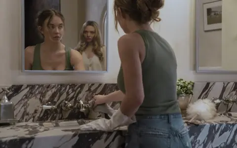 Una ragazza si pulisce il lavandino mentre un'altra la osserva riflessa nello specchio. La scena suggerisce una trama di famiglia complessa.