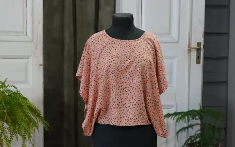 En persikofärgad blus med ett litet mönster av mörka prickar och små blommor. Blusen har en vid passform och korta, fladdermusliknande ärmar.